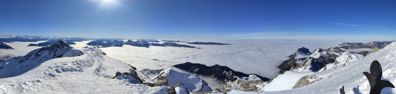 Tournette neige annecy From a high vantage point, a panoramic view of a mountainous region is seen under a clear blue sky. The sun shines brightly, casting a warm glow over the landscape. The mountains are covered in snow, with some areas showing rocky outcrops. Below, a sea of clouds blankets the valleys, creating a striking contrast between the white snow and the grey clouds. The horizon is dotted with more peaks, some of which are partially obscured by the clouds. The sky is mostly clear, with a few wispy clouds scattered about.