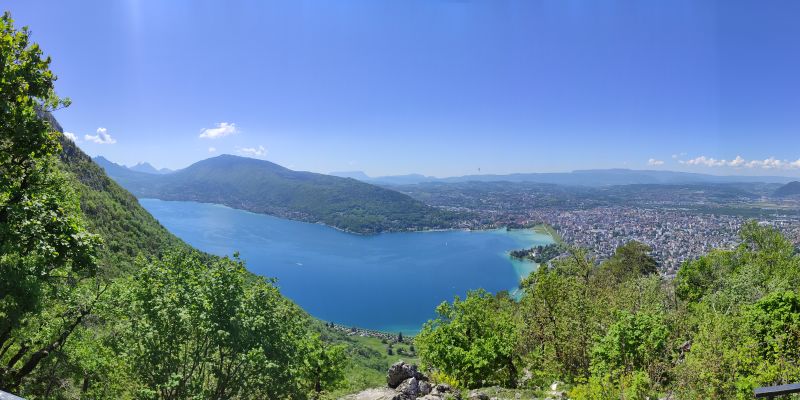 Veyrier lac annecy panoramic From a high vantage point, a panoramic view of a serene lake is visible, surrounded by lush greenery. The lake's water is calm and reflects the clear blue sky above. In the distance, a city sprawls out, with numerous buildings and structures dotting the landscape. The city is nestled between rolling hills and mountains, creating a picturesque backdrop. The sky is a vibrant blue, with a few scattered clouds adding texture to the scene. The overall atmosphere is peaceful and tranquil, with the natural beauty of the landscape taking center stage.