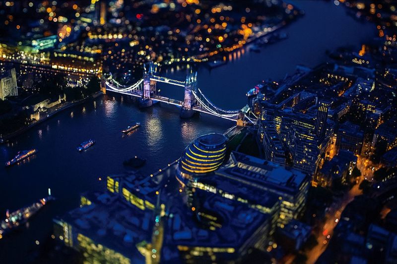 Cityscape nighttime bridge london river buildings skyline urban architecture water reflection city skyscrapers landmarks travel night aerial tower An aerial view of a city at night shows a prominent bridge spanning a river, with its towers illuminated by lights. The bridge is surrounded by water, and several boats are visible on the river. The cityscape is densely packed with buildings, many of which are brightly lit, indicating a bustling urban environment. The sky is dark, and the lights from the buildings and bridge create a warm, glowing effect. The image captures the vibrant nightlife of the city, with reflections of lights shimmering on the water's surface.