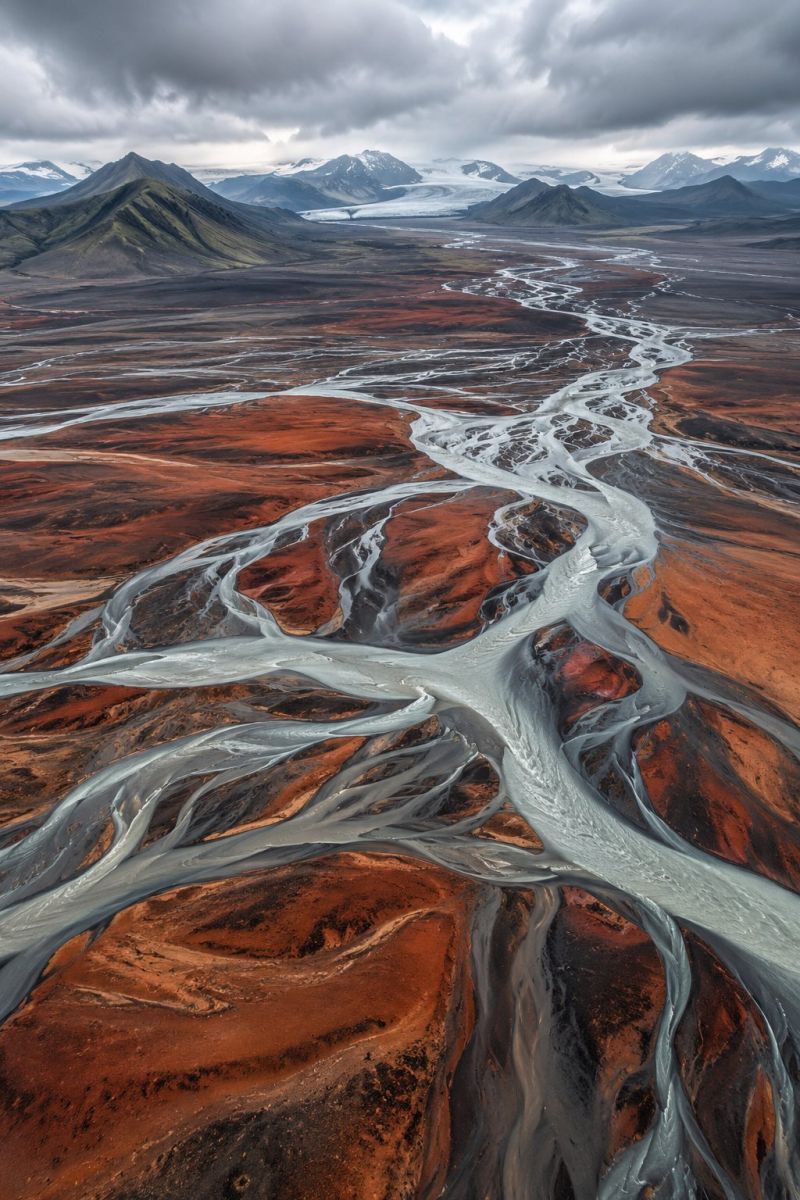 Aerial beauty colorful desert earth environment glacier glaciers ice iceland icelandic mountains river scenery wilderness An aerial view of a vast, rugged landscape dominated by a winding river that snakes through the terrain. The river is composed of multiple channels, creating a complex network of waterways. The surrounding land is a mix of earthy browns, deep reds, and patches of green, with rocky outcrops and mountains visible in the distance. The sky is overcast, casting a grayish hue over the entire scene.