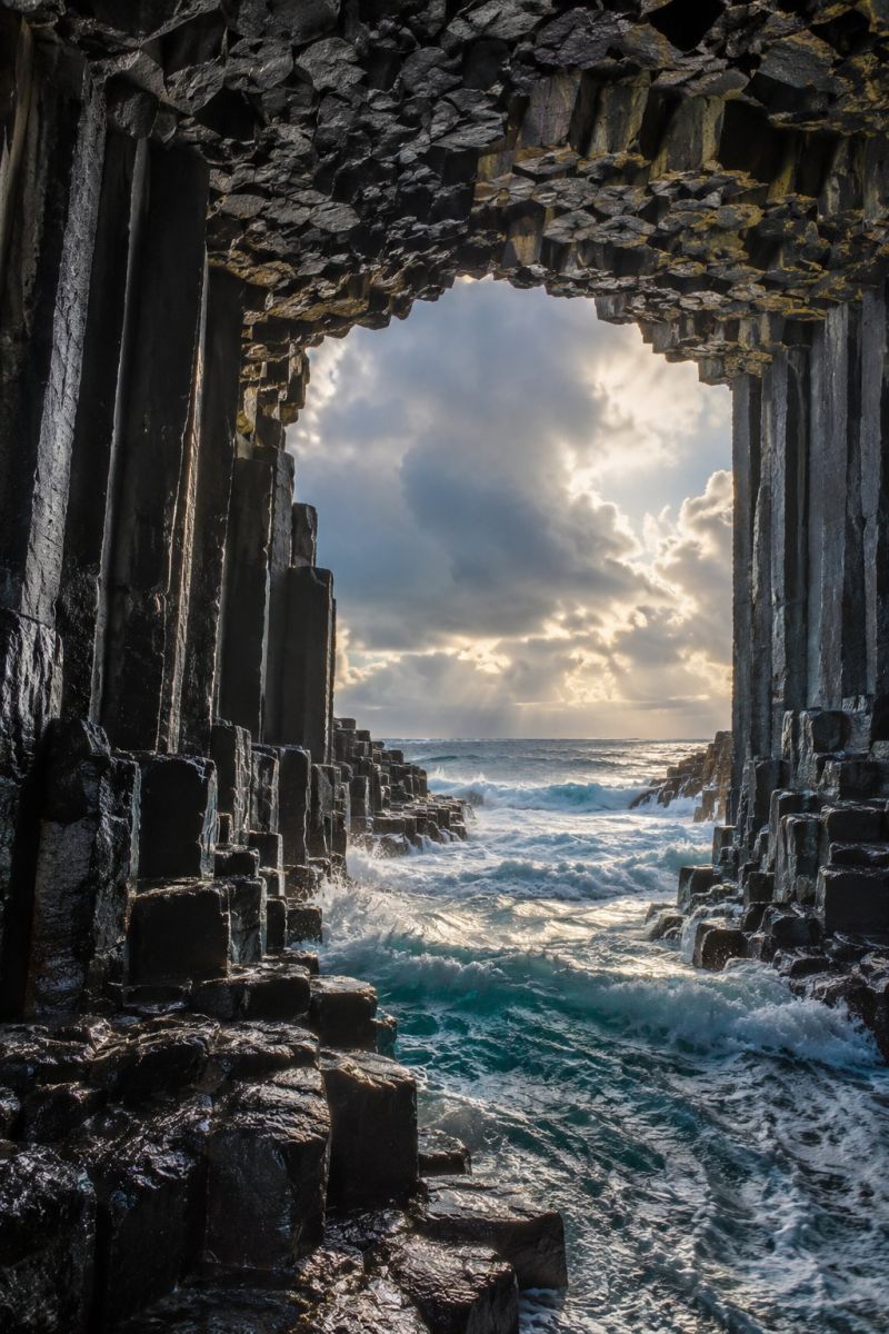 Falaise ocean eau ciel soleil vagues mer roche grotte geologie nature air marin cote erosion flanc formation geologique scène côtière spectaculaire avec une grande colonne de basalte hexagonale. Les colonnes sont sombres et dentelées et s'élèvent verticalement depuis la mer. Le haut des colonnes forme une arche naturelle, à travers laquelle la lumière du soleil traverse et illumine l'intérieur. La mer est agitée, les vagues s'écrasant contre la base des colonnes, créant une mousse blanche mousseuse. Le ciel au-dessus est partiellement nuageux, avec des taches de nuages bleus et blancs. La scène d'ensemble est spectaculaire et impressionnante, mettant en valeur la puissance de la nature et la beauté des formations géologiques.