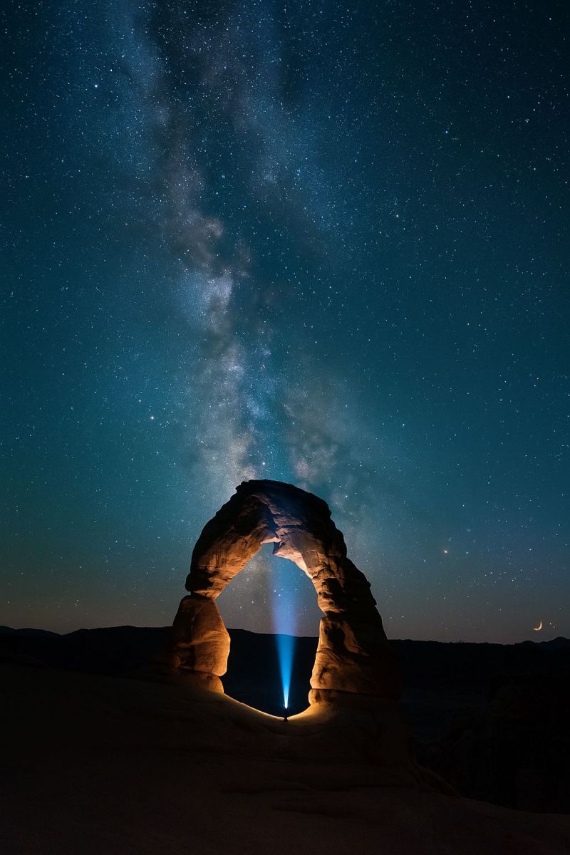 Nuit ciel etoiles obscurite arc roche nature air desert lune galaxie astronomie astrophotographie voyage aventure exploration voie lactee Une formation rocheuse naturelle en forme d'arche est mise en évidence sous un ciel nocturne rempli d'étoiles. L'arche est éclairée par une lumière vive, possiblement une lampe de poche, créant un effet dramatique. Le faisceau lumineux traverse l'arche, projetant une longue ombre sur le sol. Le ciel est d'un bleu profond, parsemé d'innombrables étoiles, et la Voie Lactée est faiblement visible, ajoutant à l'ambiance céleste. L'arche est faite de roche rouge-brun, et la zone environnante semble être un paysage désertique, avec le sol recouvert de petits cailloux et de sable. La scène globale est sereine et impressionnante, capturant la beauté de la nature la nuit.