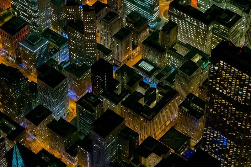 Urbain nuit ligne horizon batiments architecture moderne gratte ciel aerien ville centre affaire financier quartier tours verre reflets Une vue aérienne d'une ville animée la nuit, montrant un paysage urbain dense rempli de grands bâtiments. Les bâtiments varient en taille et en design, certains présentant des styles architecturaux modernes et d'autres apparaissant plus traditionnels. Beaucoup de bâtiments sont illuminés par des lumières, créant un paysage urbain vibrant et dynamique. Les rues en dessous sont étroites et entrecroisées, avec certaines zones apparaissant plus congestionnées que d'autres. L'atmosphère générale est celle de l'activité et de l'énergie, avec la ville vivante et prospère même dans l'obscurité de la nuit.